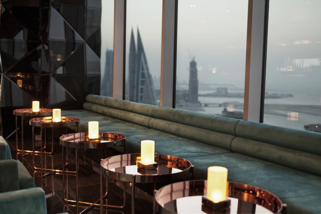 The Domain Bahrain Hotel And Spa - Adults Friendly 16 Years Plus 마나마 외부 사진 The photo depicts a stylish lounge area with a modern aesthetic. There are several circular tables made of metallic materials, each topped with a small glowing candle. A long, plush seating area in a teal color runs along the window, offering a view 